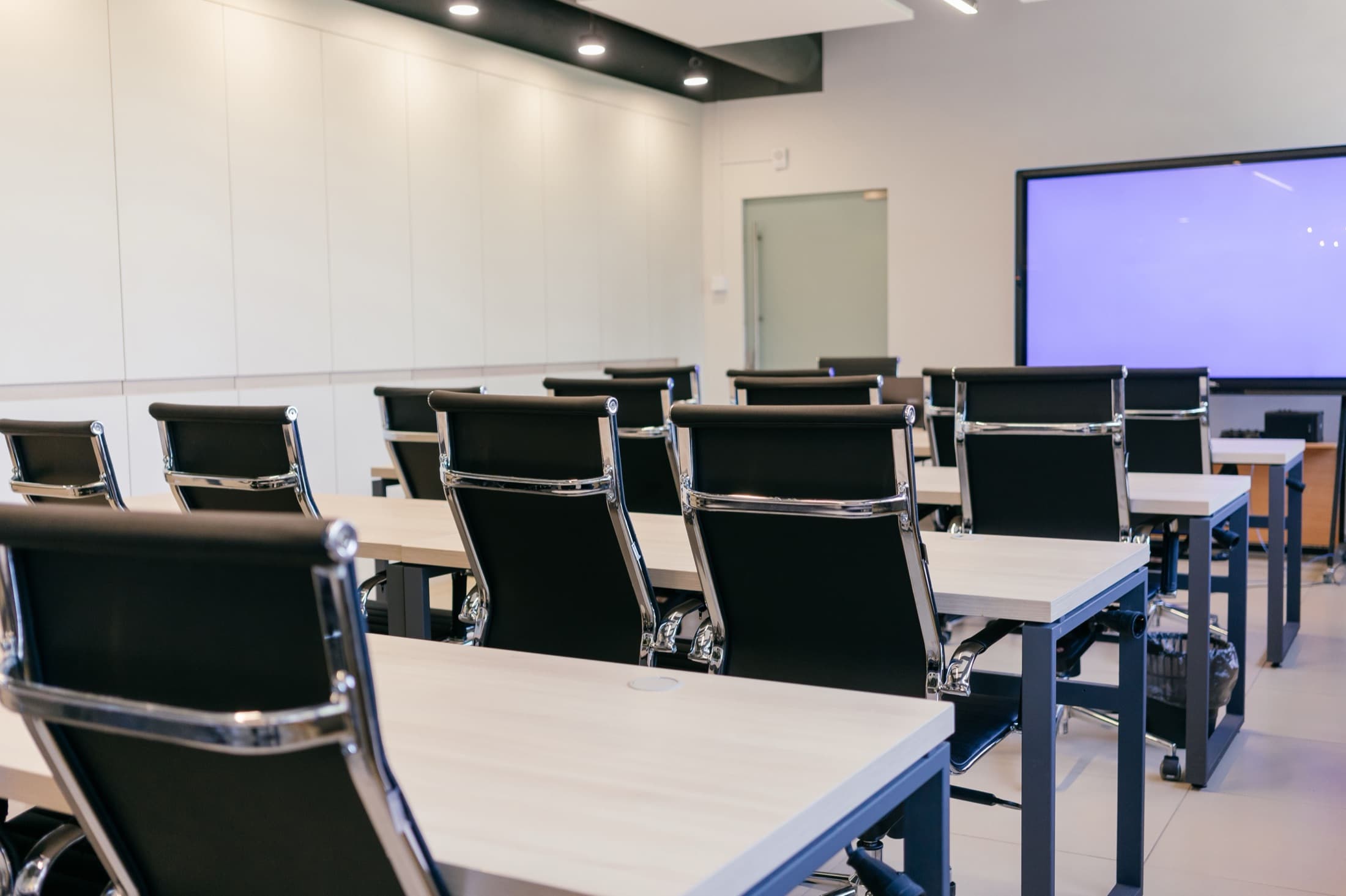An empty seminar room, desks and chairs in rows facing a display screen.